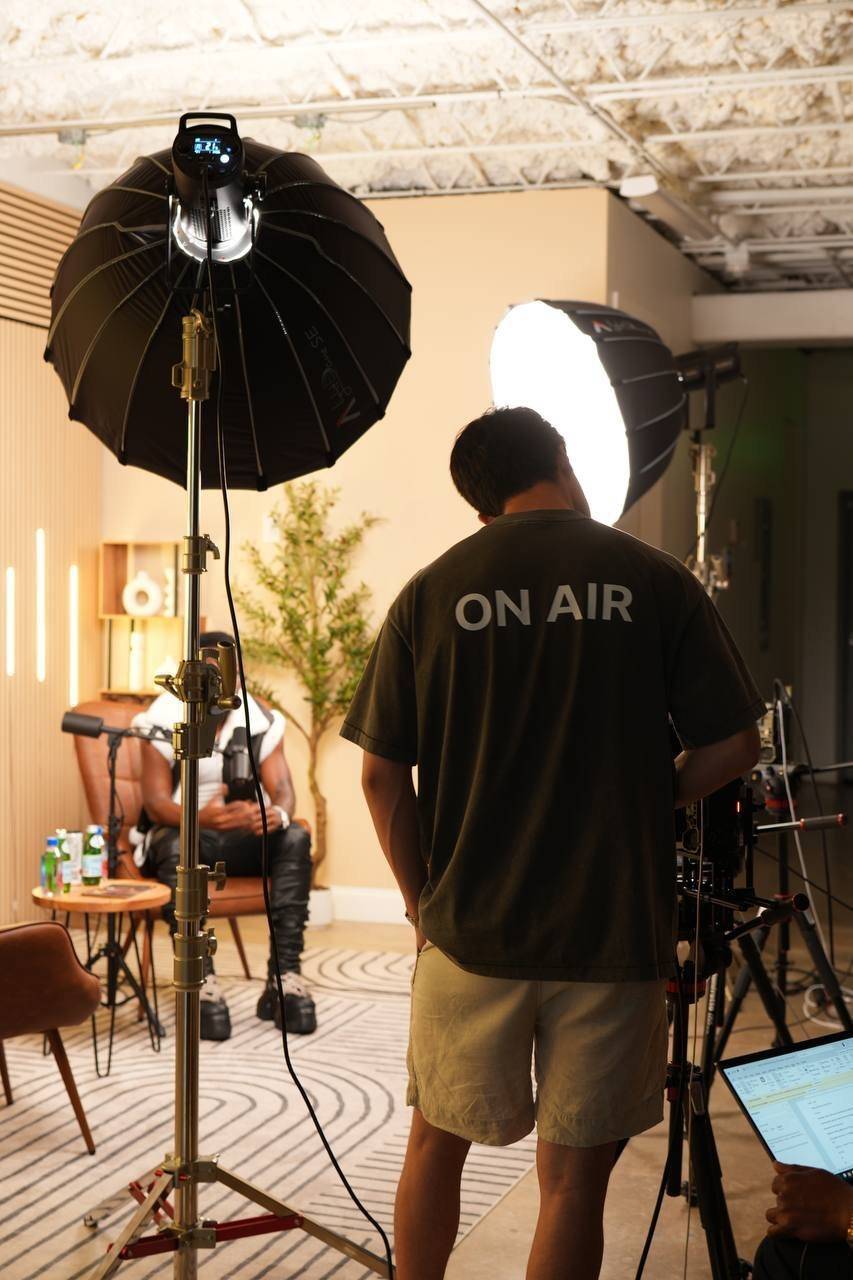 A person in an ON AIR t-shirt stands at a camera rig between two large studio lights, with a seated person in an armchair in the background