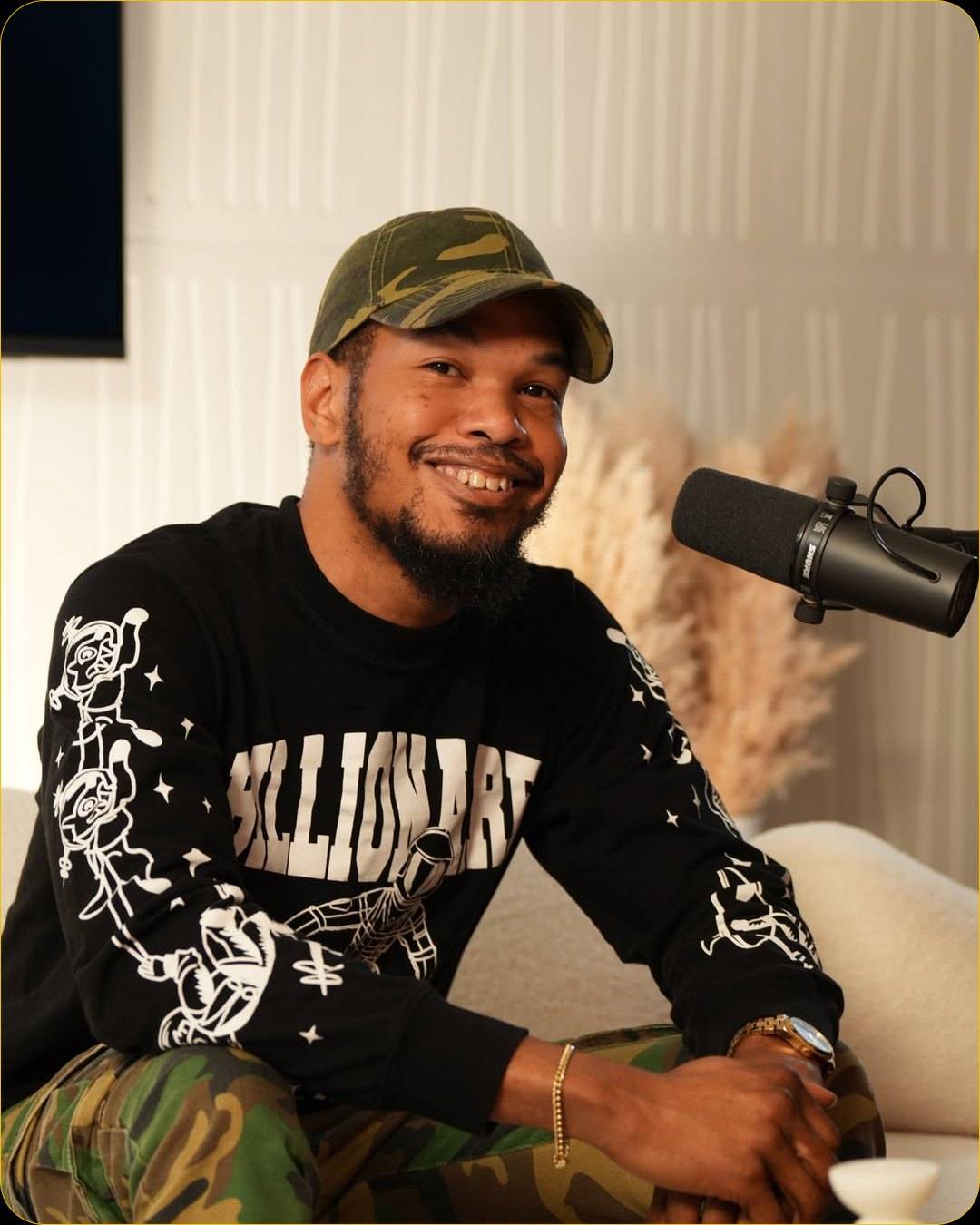 A smiling man in a camouflage cap and a black graphic sweatshirt sits on the white sofa beside a microphone, with pampas grass behind him