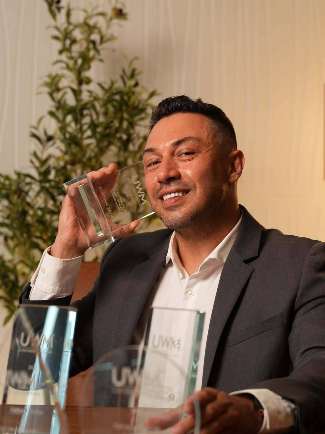 A man in a dark blazer smiles and holds a glass plaque beside his face, with two more glass plaques on the wooden table in front of him and olive branches behind