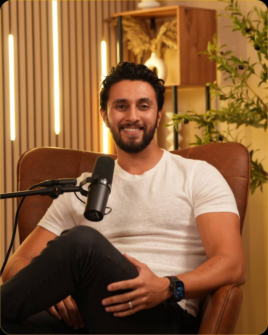 A smiling man in a white t-shirt and black jeans sits back in a leather armchair, a microphone on a boom arm beside him, with wooden shelves and olive branches behind