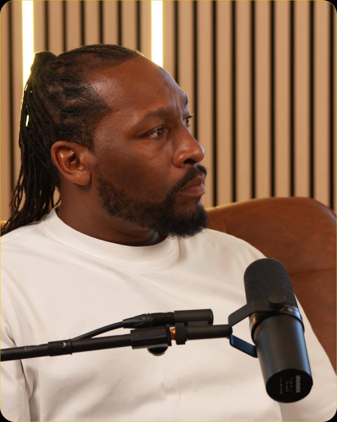 A man in a white sweatshirt looks away from the camera in a leather armchair, with a microphone on a boom arm in the foreground and wood slats with light bars behind him