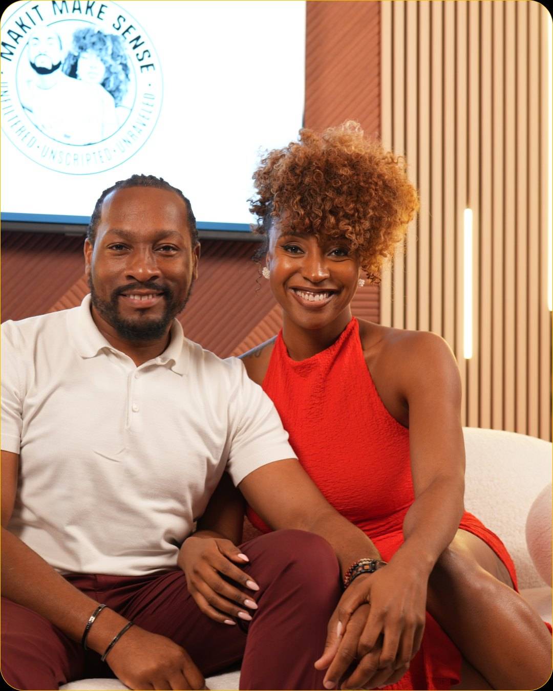 Two guests sit close together on the sofa and smile at the camera, with a round podcast logo on the display and wood slats behind them