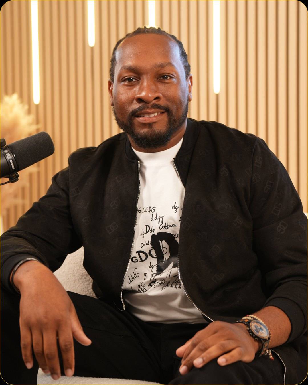 A smiling man in a black bomber jacket over a graphic white t-shirt sits on the sofa next to a microphone, with pampas grass and light bars behind