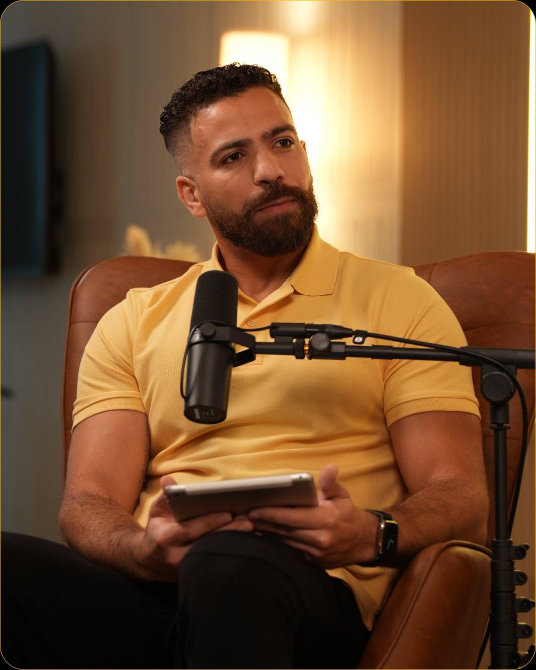 A man in a yellow polo holding a tablet at the microphone on a leather armchair in Studio #1