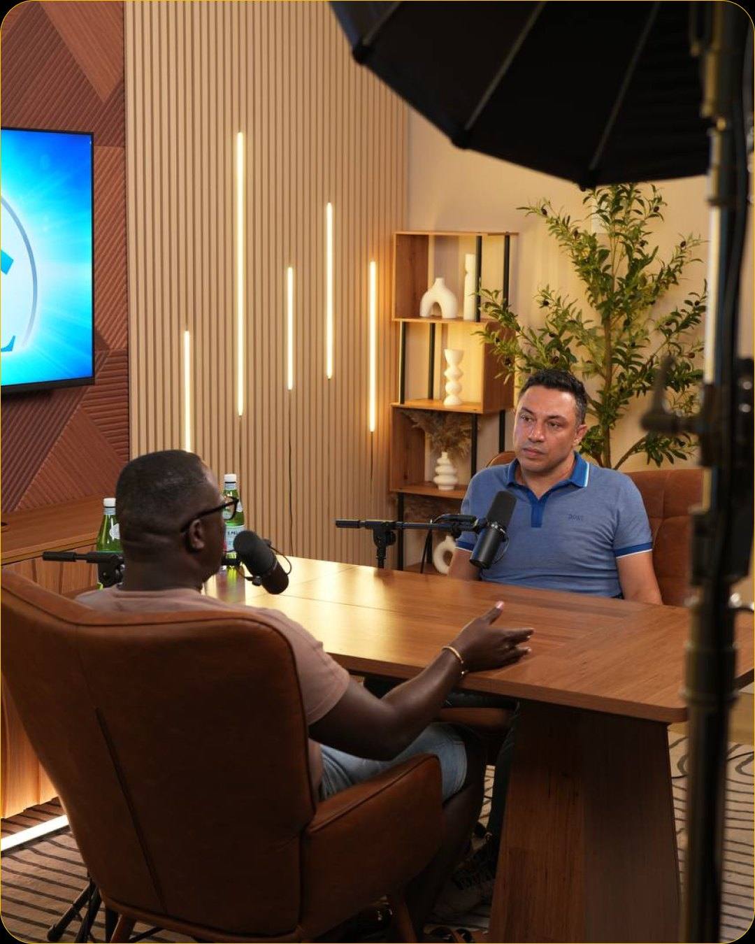 Two people recording at the wooden table of Studio #4, with a wall display and LED light bars behind them