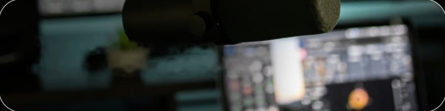 A studio microphone in the foreground, with a small plant and a monitor showing software out of focus behind it