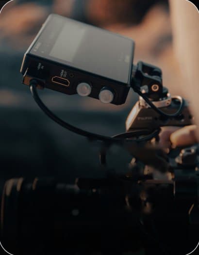 Close-up of a camera monitor and cable on a camera rig, with a hand on the rig and the lens out of focus below
